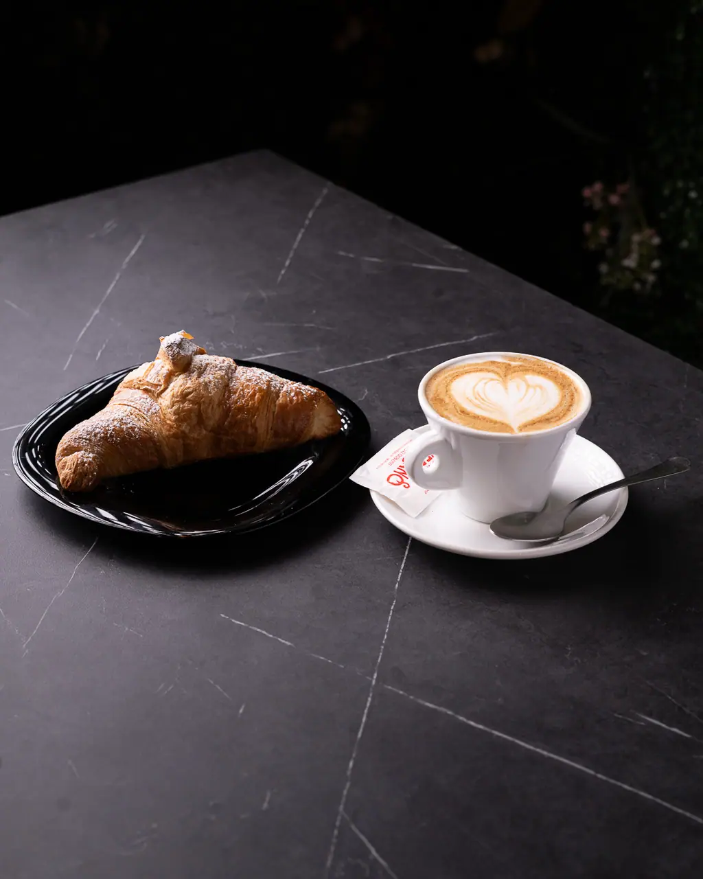 Cappuccino con latte art a cuore e cornetto su tavolo scuro